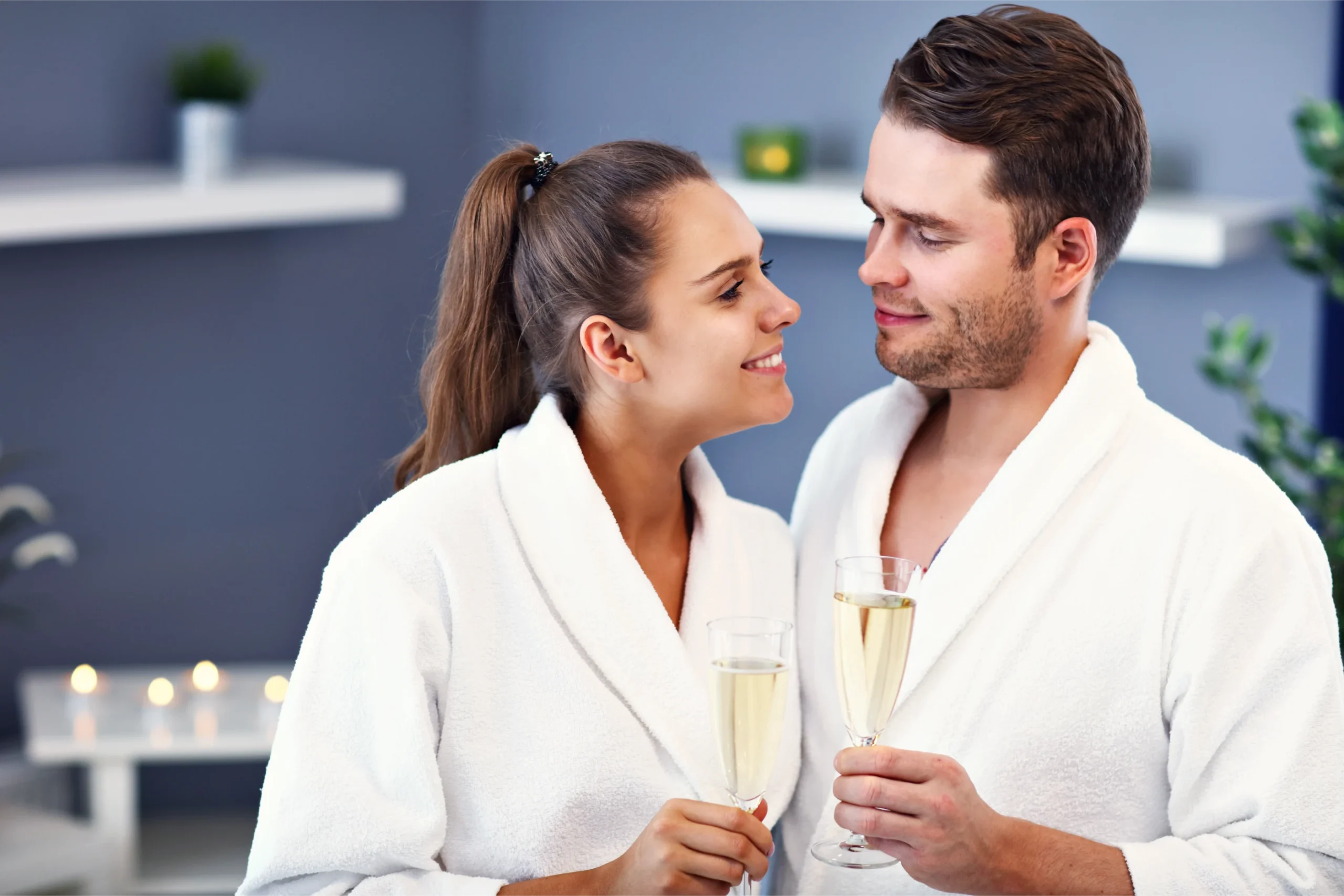 Happy couple in bathrobes holding champagne flutes, looking at each other and smiling in a spa setting.