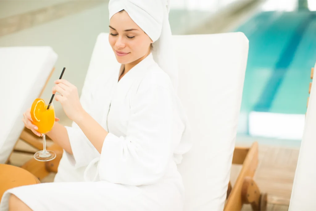 Young woman in a white bathrobe and head towel sips orange juice, relaxing on a lounge chair by a spa pool.